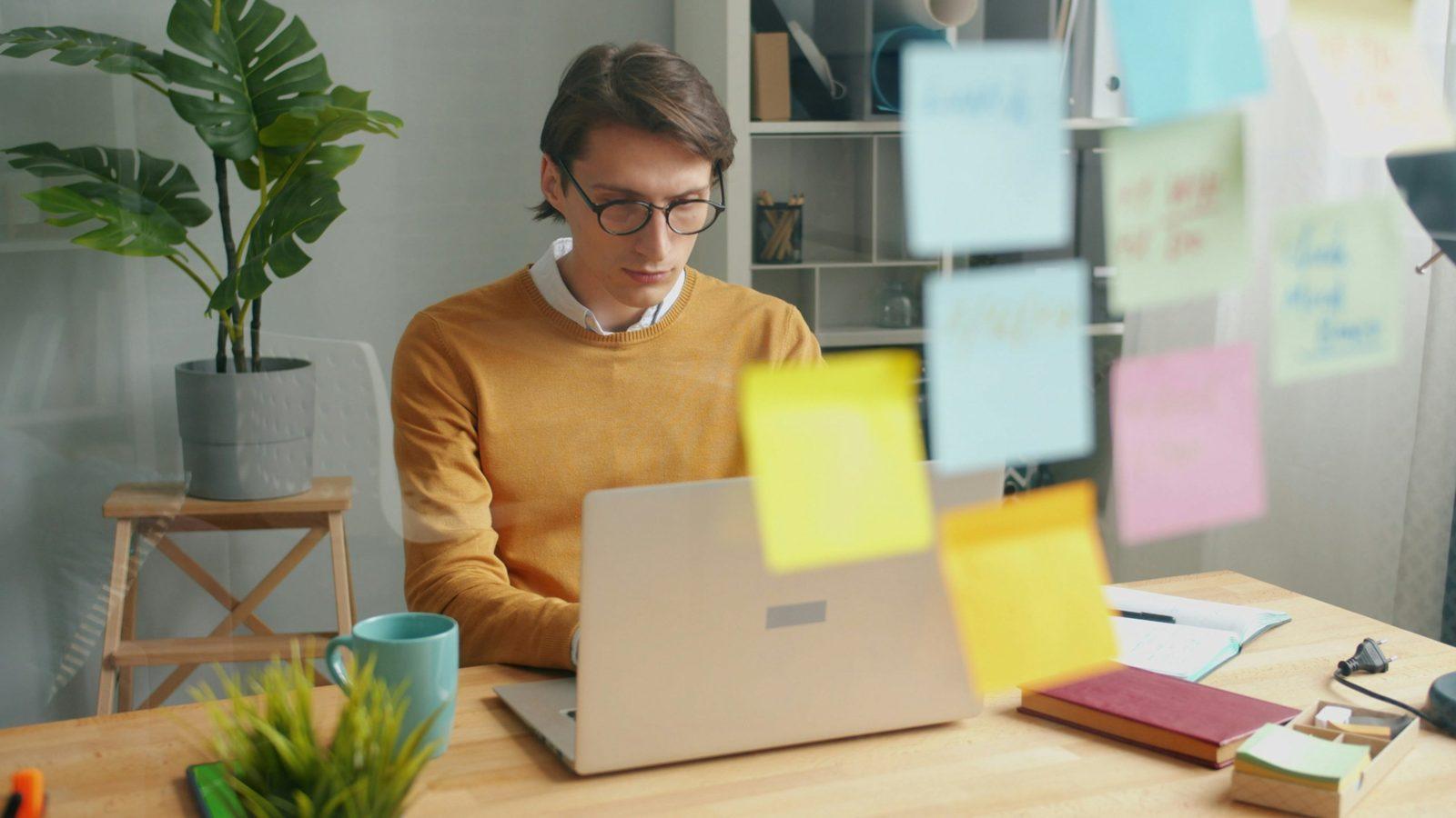 A man working on a laptop at his desk, with sticky notes on a glass wall in front